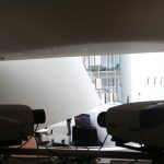 Bir sergi salonunda yan yana duran iki projeksiyon cihazının önden görünümü, arka planda projeksiyon perdesine yansıyan görüntü ve cam cepheli bir bina