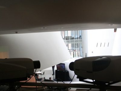 Bir sergi salonunda yan yana duran iki projeksiyon cihazının önden görünümü, arka planda projeksiyon perdesine yansıyan görüntü ve cam cepheli bir bina