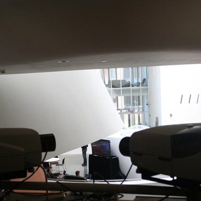 Bir sergi salonunda yan yana duran iki projeksiyon cihazının önden görünümü, arka planda projeksiyon perdesine yansıyan görüntü ve cam cepheli bir bina